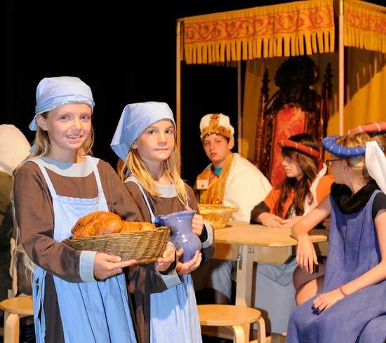 Kinder in mittelalterlichen Kostümen bei einem Theaterstück mit Essensrequisiten und Thron im Hintergrund